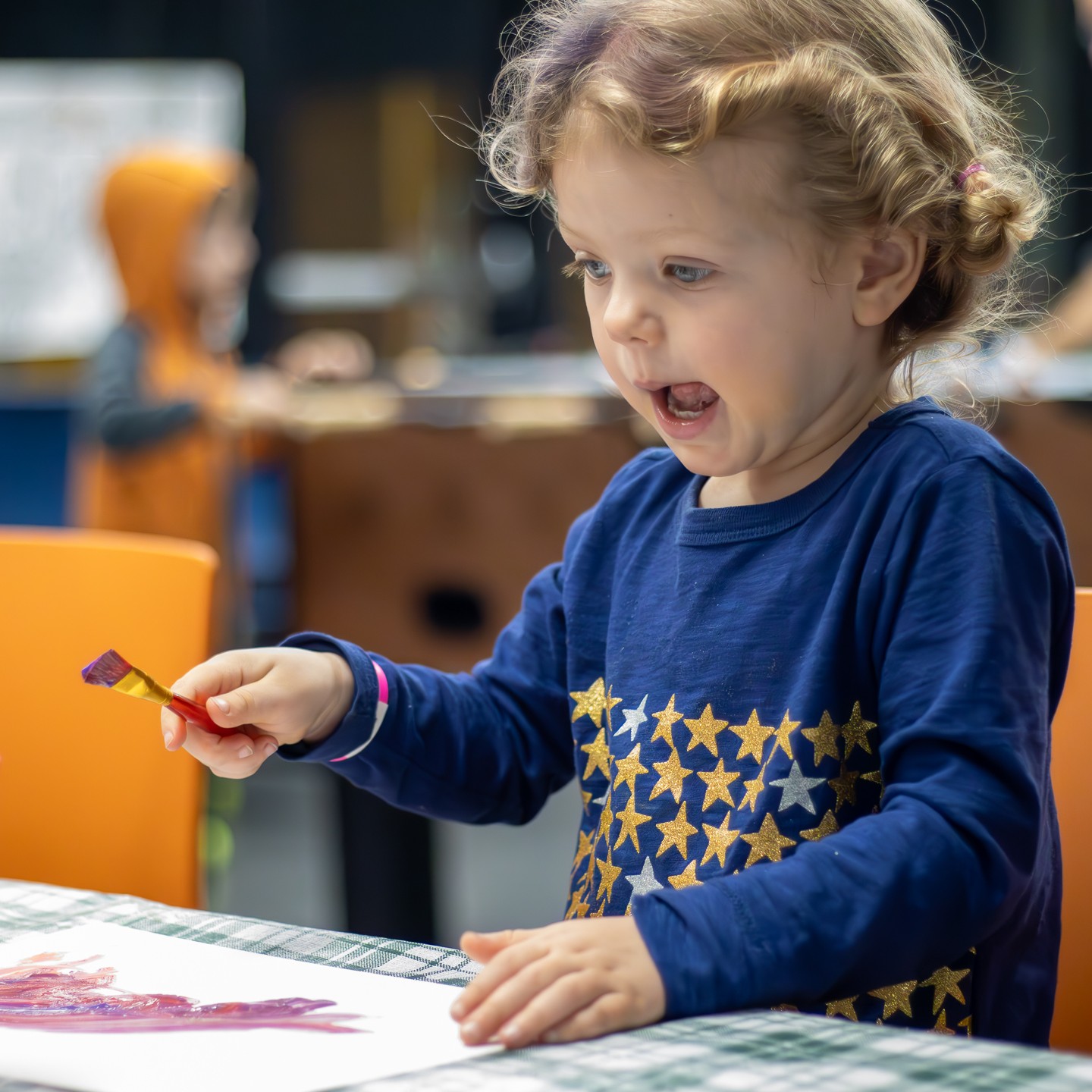 an excited child holding a paint brush over a white paper with red paint on it